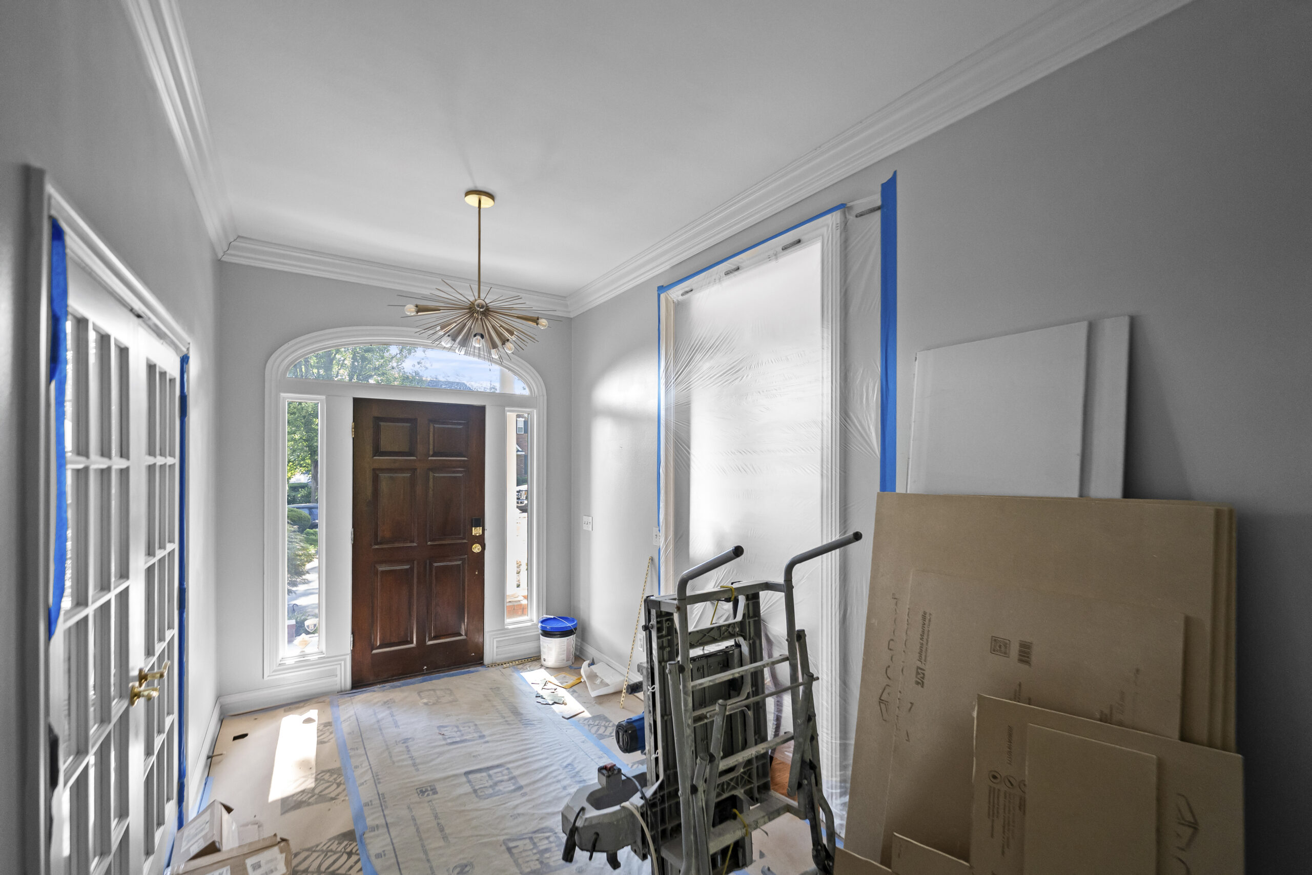 Home entryway mid-renovation with painter's tape on trim, floor protection paper, and drywall panels stacked against the wall