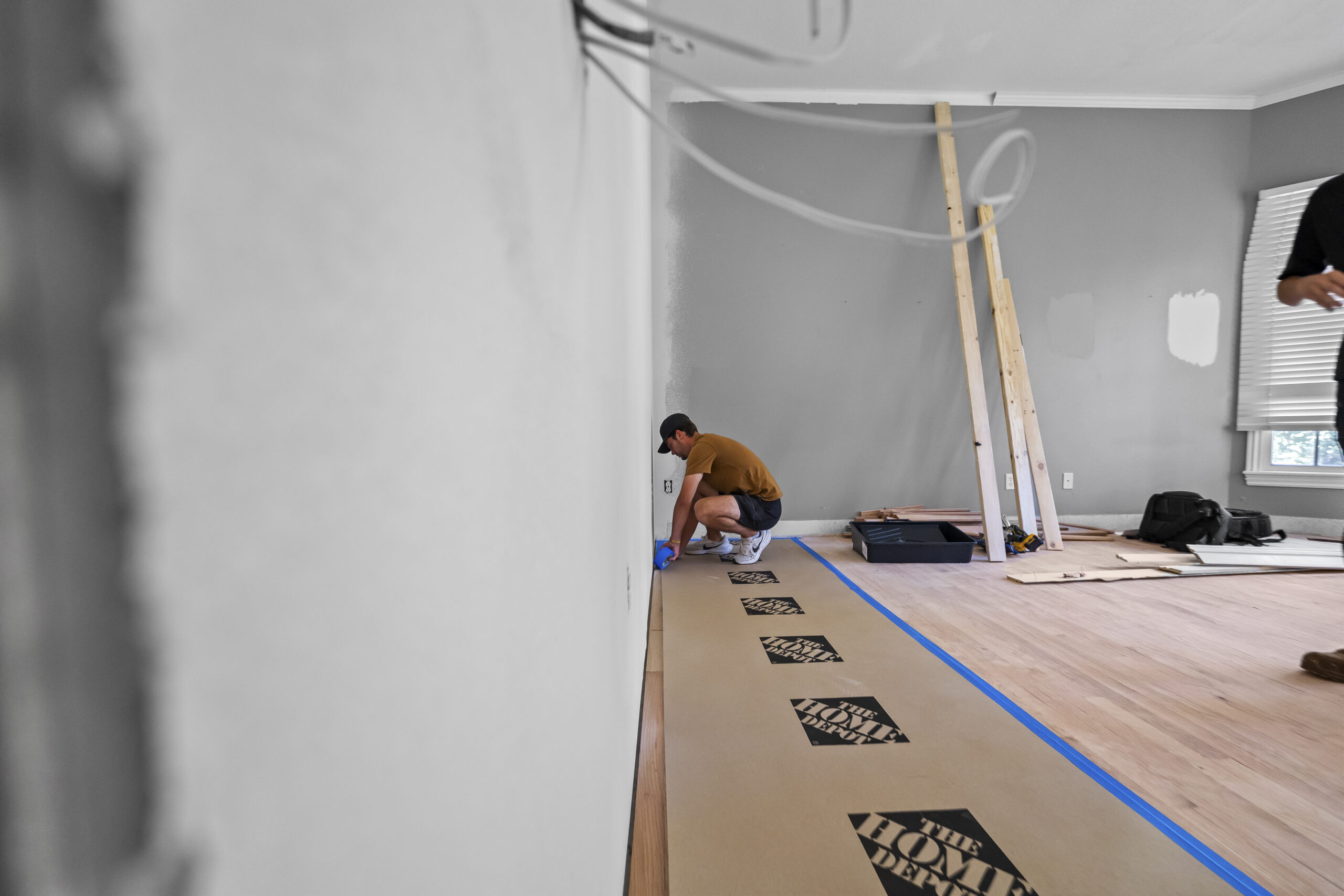 Contractor kneeling to install hardwood flooring in a commercial space with floor protection board and a ladder nearby